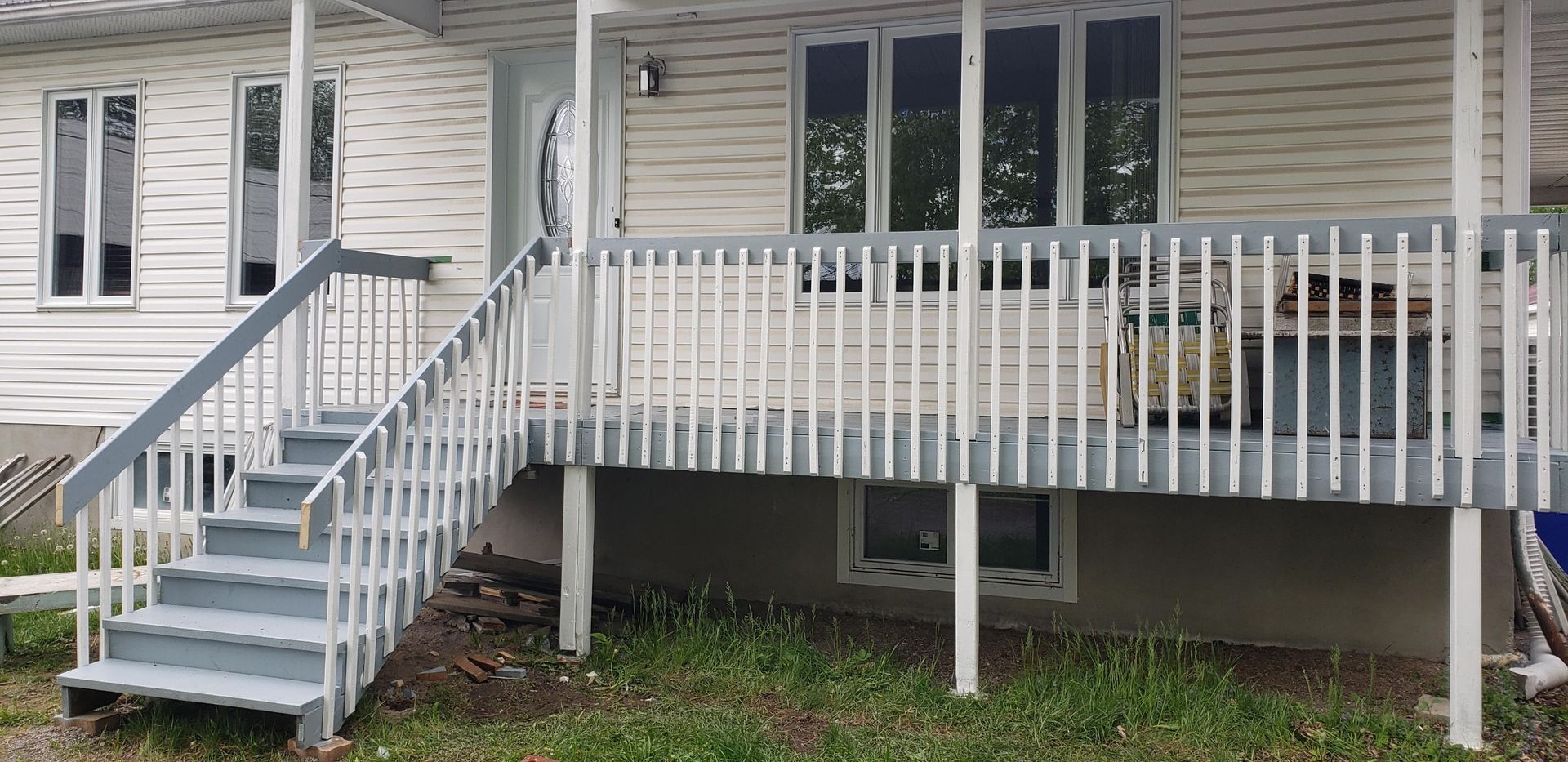 A raised wooden porch with grey stairs and railing attached to a house with beige vinyl siding and white trim.