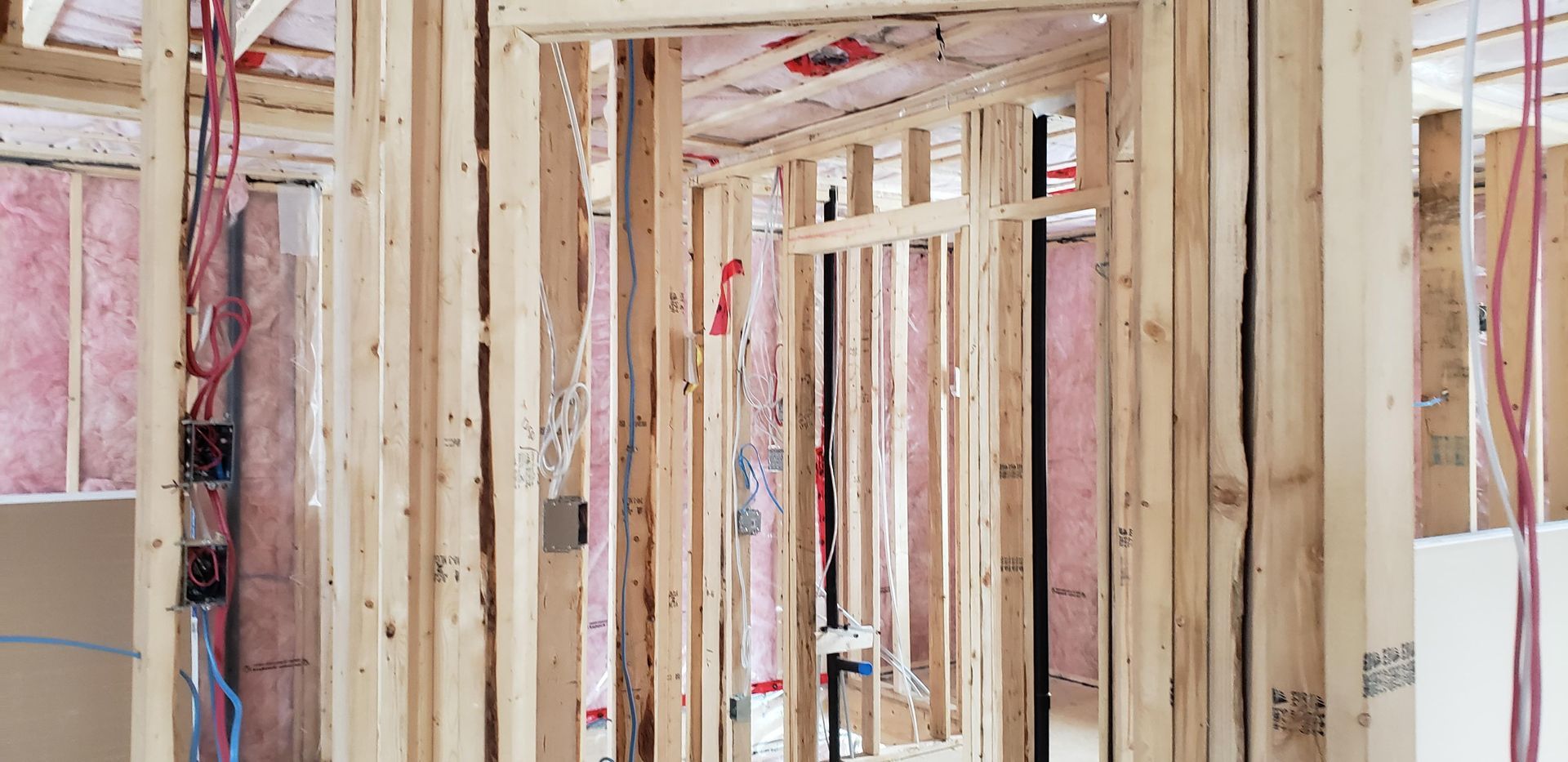 Un chantier de construction montrant une ossature murale en bois, une isolation en fibre de verre rose et des câbles électriques apparents.