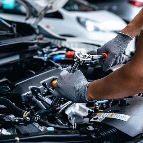 A person wearing grey work gloves uses a metal ratchet tool to repair a car engine in a garage.