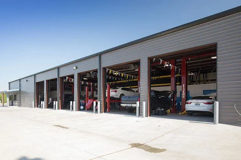 An exterior view of a modern automotive repair shop with five open service bays, showing vehicles undergoing maintenance.