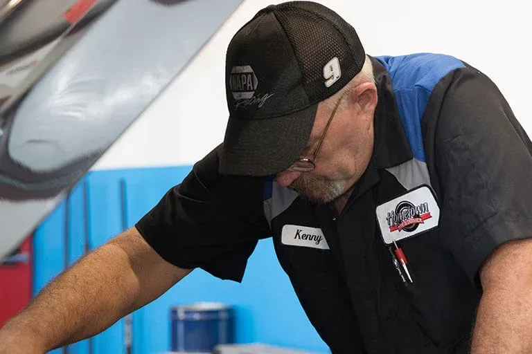 A technician in a black and blue uniform and a NAPA baseball cap leans over an open car hood in a workshop.