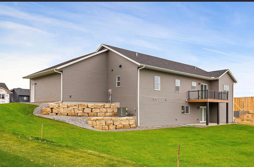 A gray, two-story suburban home with a back deck, stone retaining walls, and a large grassy yard under a blue sky.