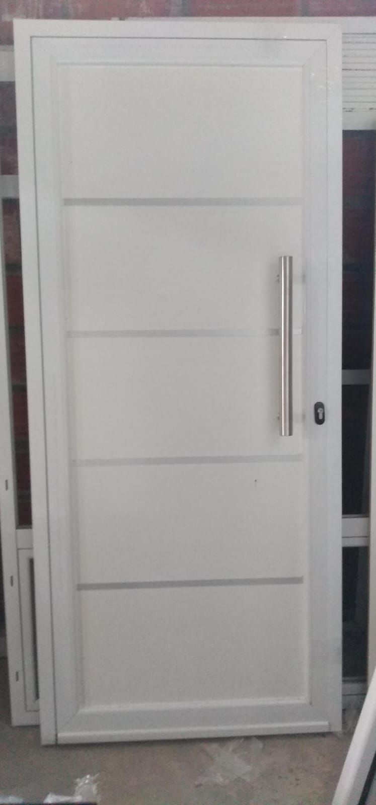 Puerta de aluminio blanco con paneles horizontales, manija vertical plateada y cerradura, sobre un fondo interior.