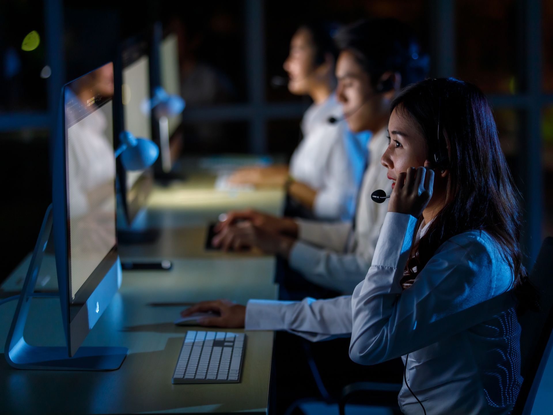 Three people wearing headsets working at computers in a dimly lit office.