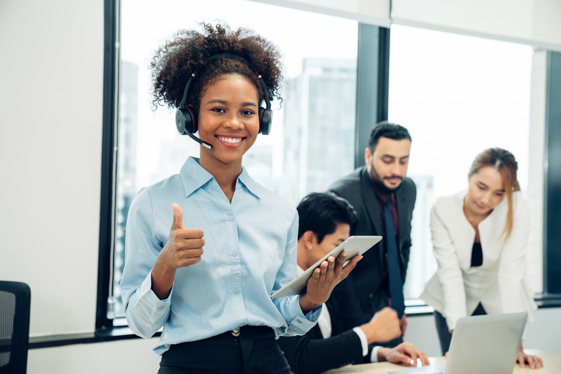 Woman with headset gives thumbs up, smiling in office; colleagues in background.
