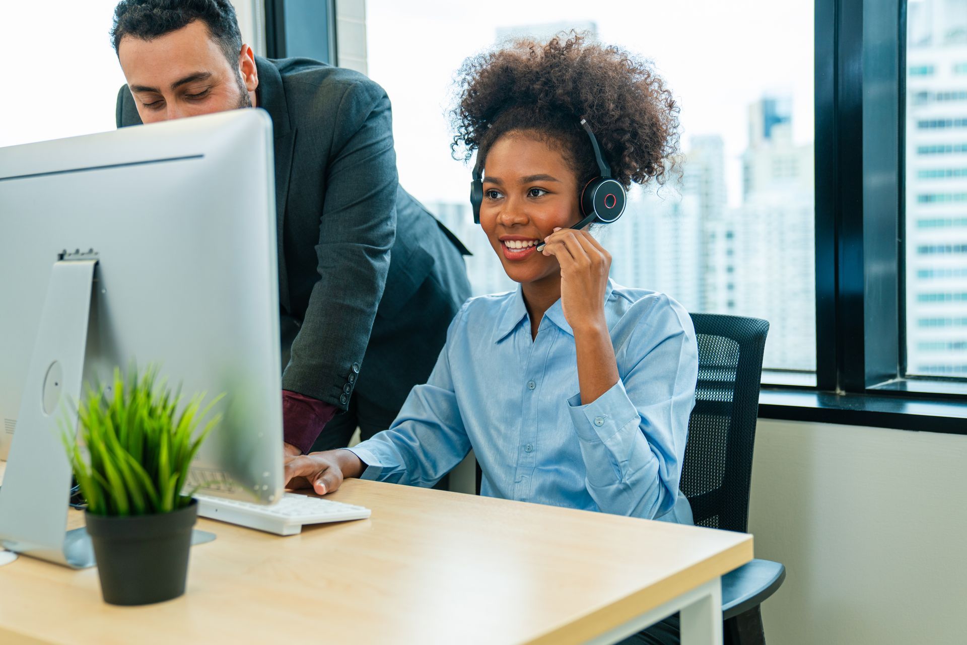 Woman in headset smiles at a computer, assisted by a person leaning over her shoulder in an office setting.