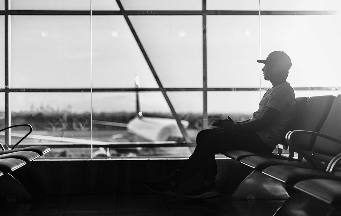Young man traveling with cremains, sitting at an airport gate looking out a big glass window