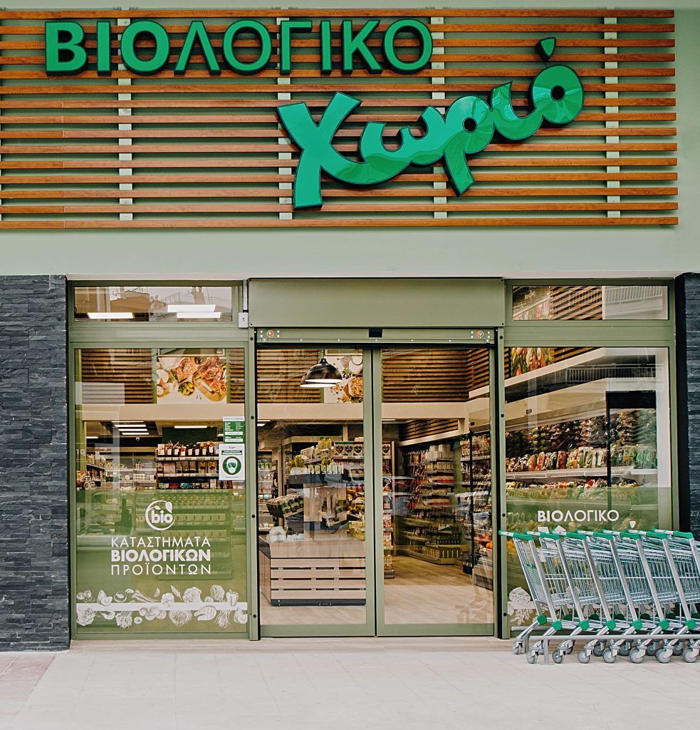 Exterior of a Greek organic grocery store: green sign, glass doors, shopping carts, and a wooden facade.