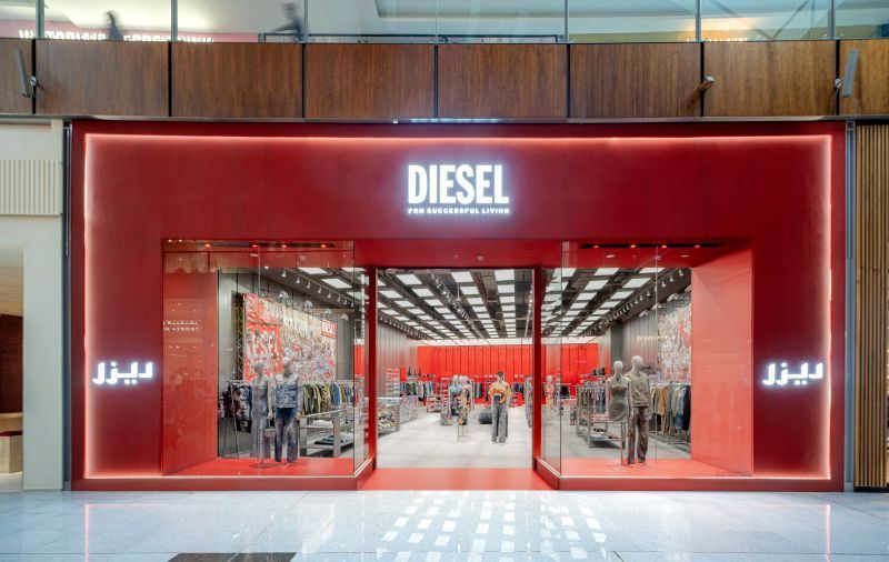Diesel store exterior: red facade, glass doors, mannequins, interior displays, and Arabic writing on the right.
