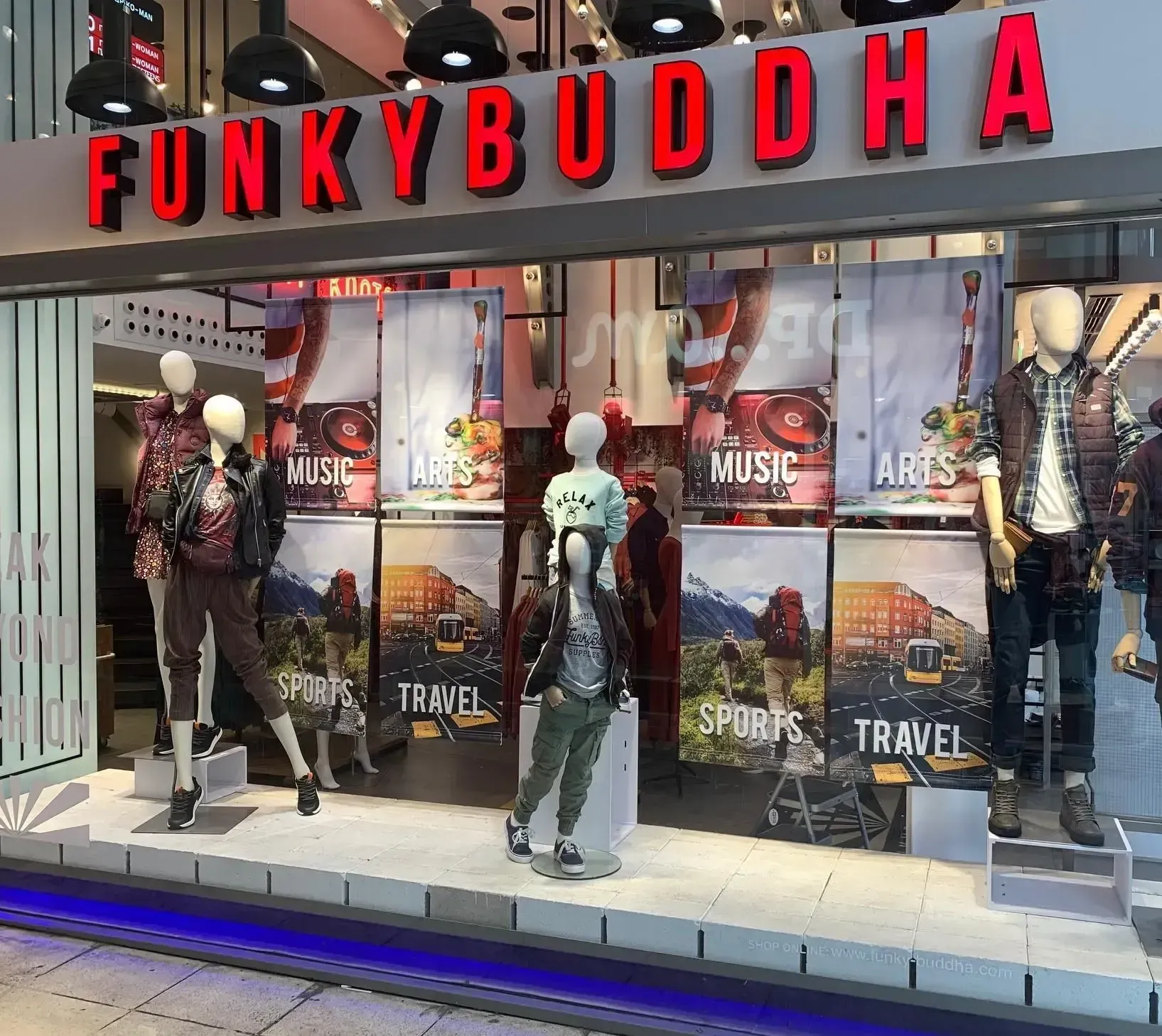 Funky Buddha store front with mannequins displaying clothing, posters with travel themes. Red letters on grey background.