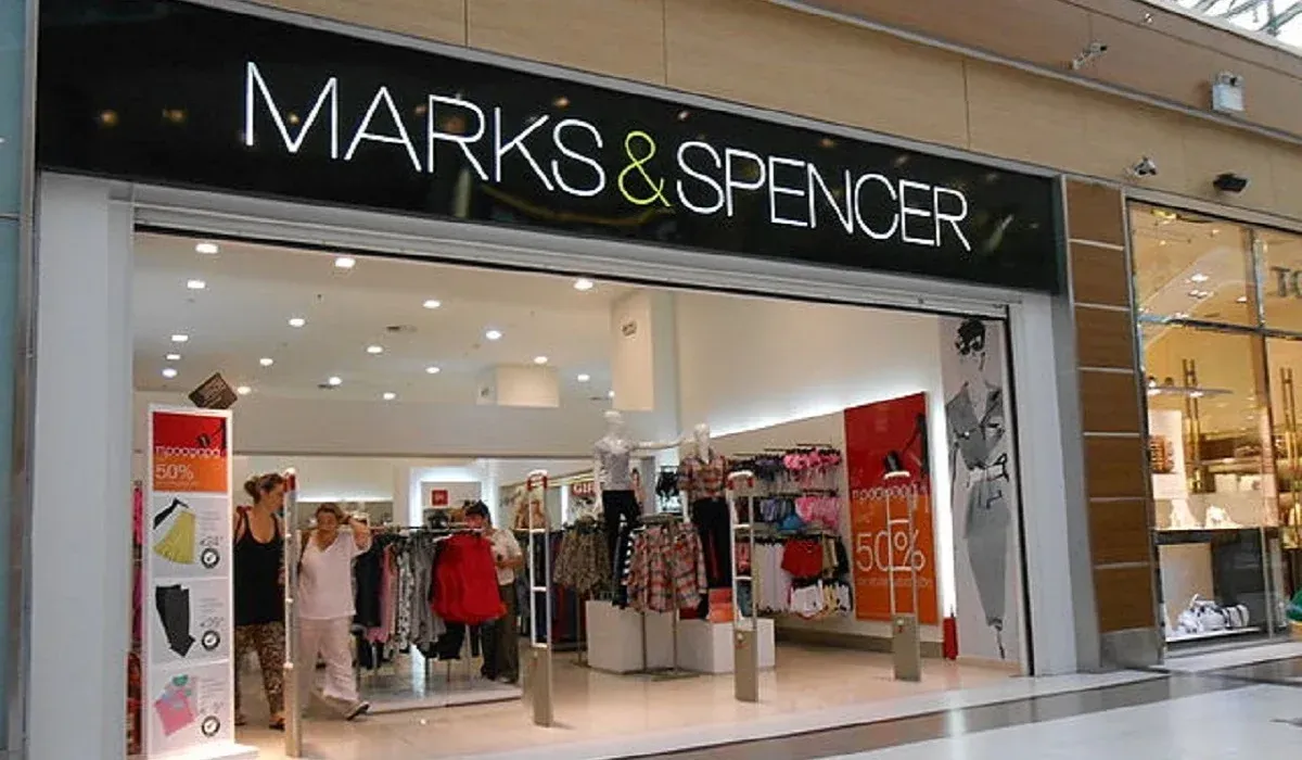 Marks & Spencer store front with clothing displays, light sign. Two people inside.