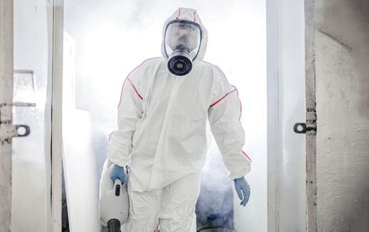 Persona con tuta protettiva e respiratore che spruzza disinfettante su una porta, in mezzo alla nebbia.