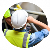 Operaio edile che indossa un casco e un giubbotto di sicurezza e guarda il telefono mentre è in piedi vicino a un'impalcatura.