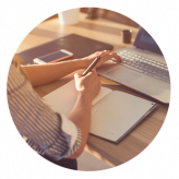 Persona che scrive su un quaderno accanto a un computer portatile e uno smartphone su una scrivania di legno.