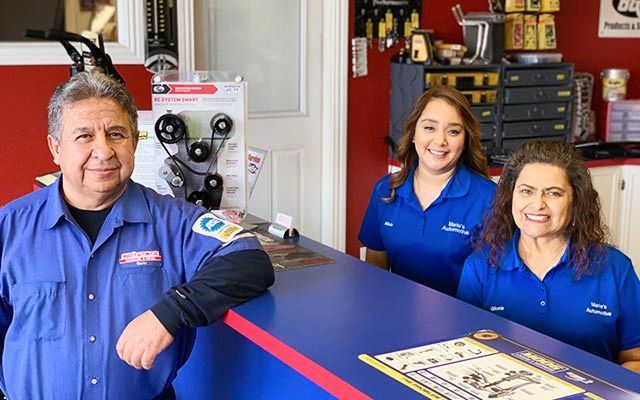 Three mechanics in blue uniforms stand by a workbench in an auto shop, smiling at the camera. | Mario’s Automotive