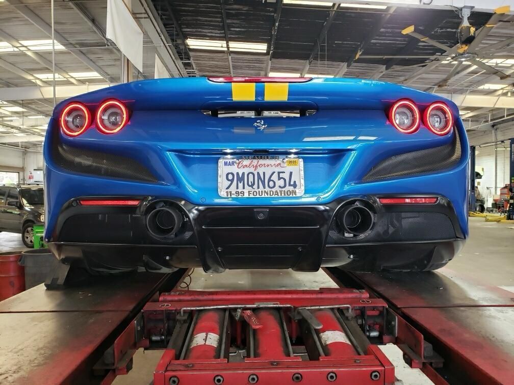 Blue Ferrari on a lift, rear view. California license plate, yellow/gold stripe detail. Exhaust visible.