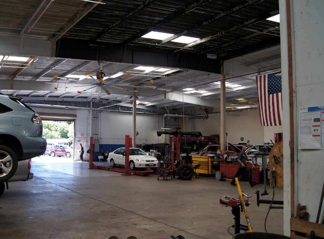 Inside a vehicle repair shop, cars on lifts, tools, American flag, ceiling with skylights, open garage door.