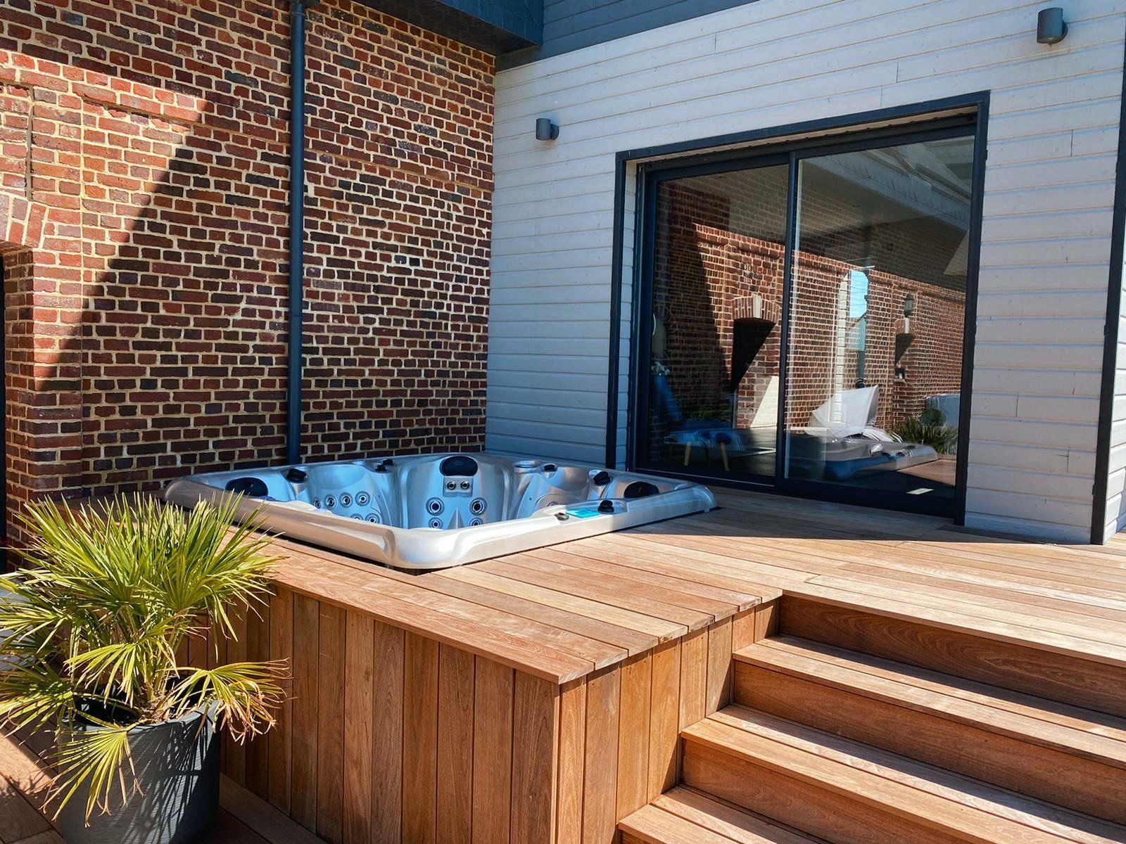Un bain à remous est installé sur une terrasse en bois à côté d'un bâtiment en briques.