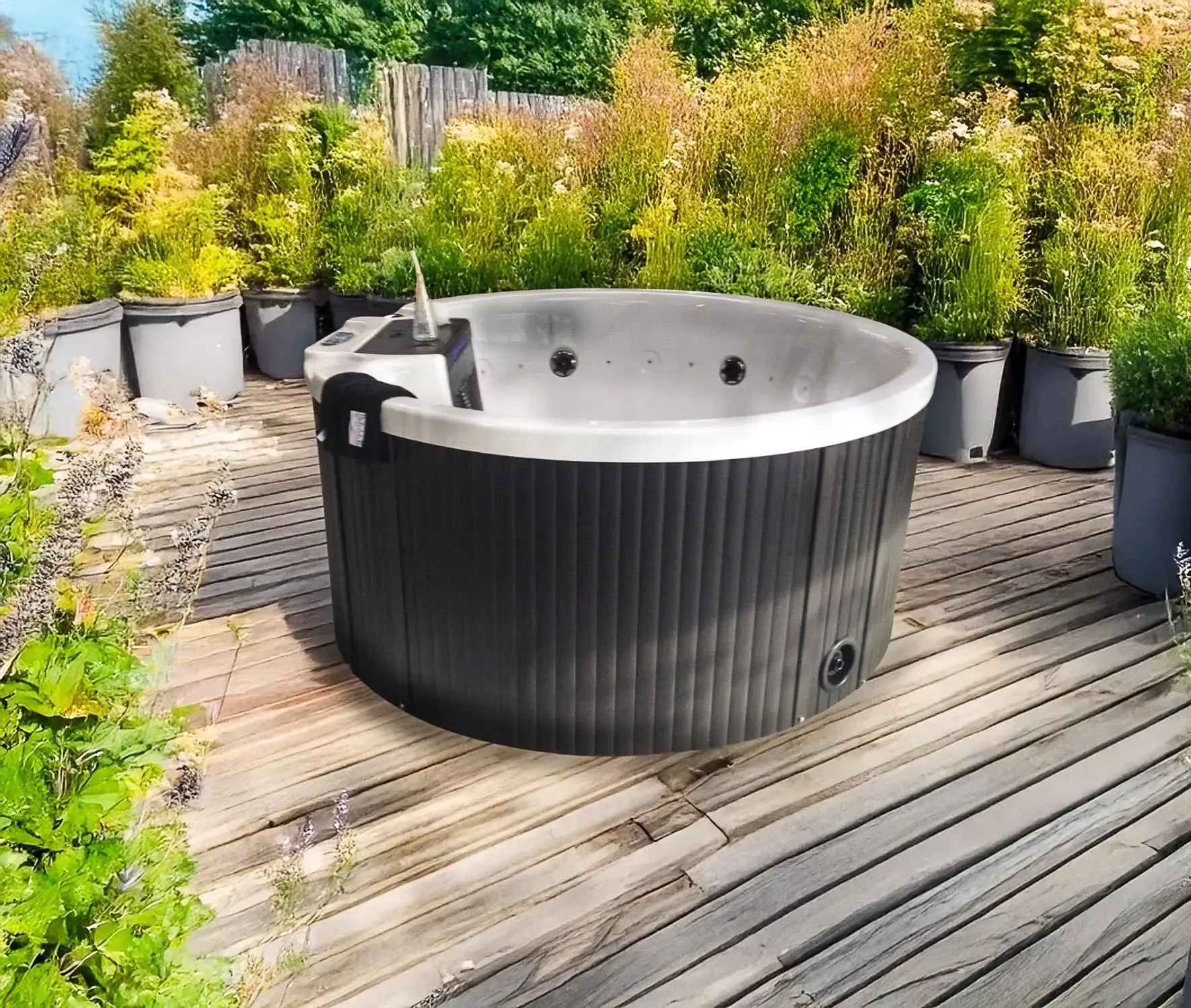 Un jacuzzi noir sur une terrasse en bois, entouré de plantes en pot et de feuillage. Cadre extérieur ensoleillé.