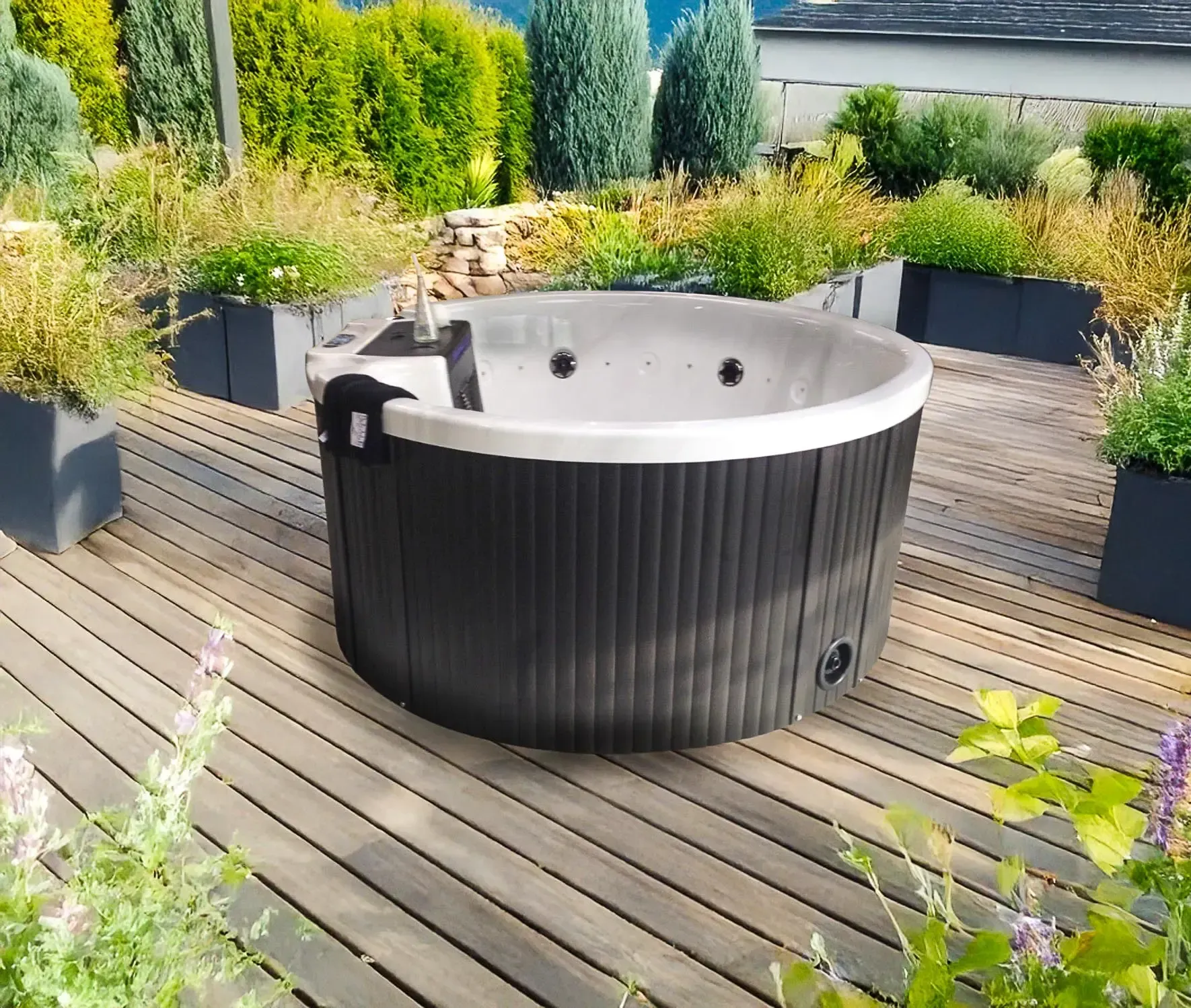 Un jacuzzi rond est installé sur une terrasse en bois, entourée de plantes en pot et de verdure. Son extérieur est sombre et son rebord intérieur est blanc.