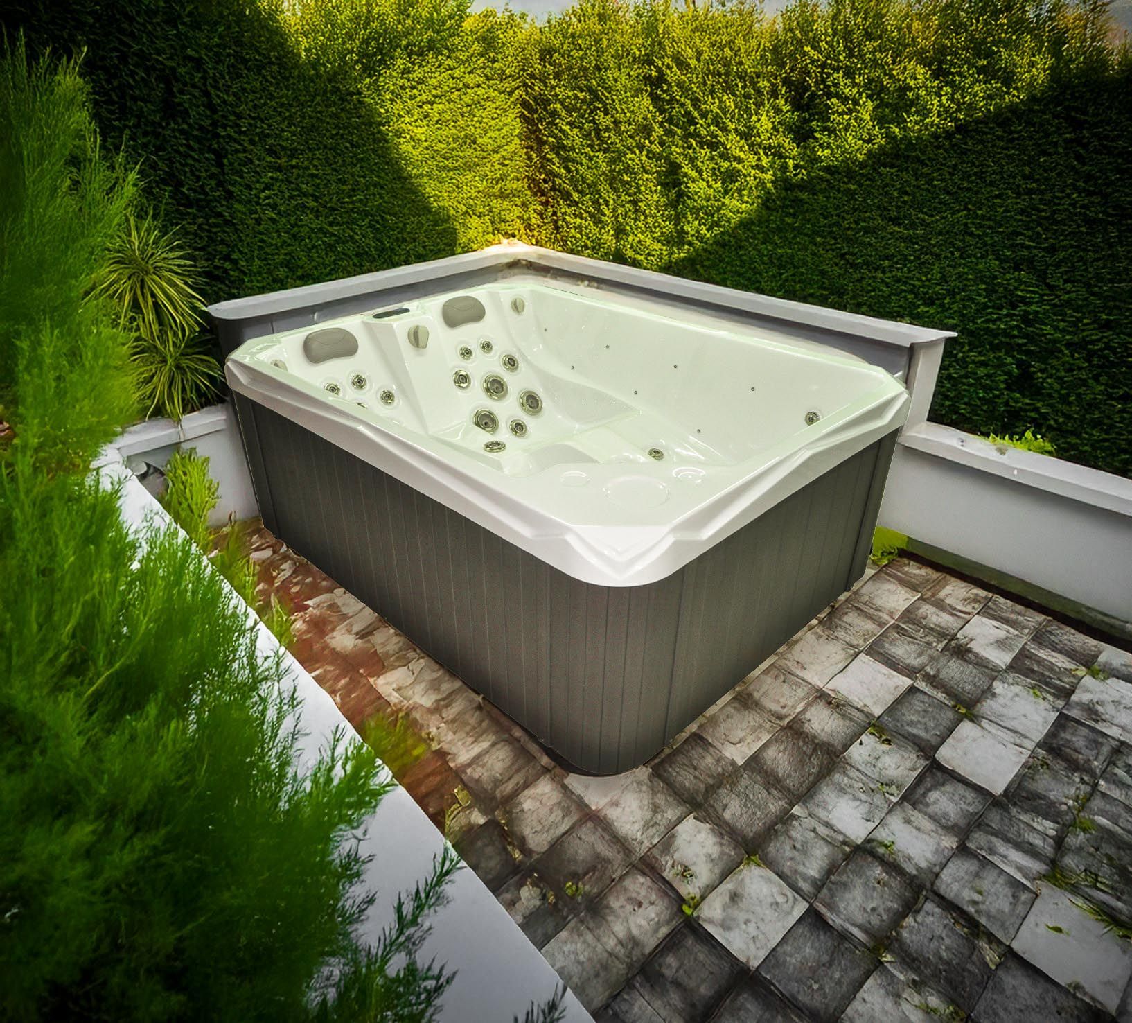 Spa installé sur une terrasse en pierre, entouré de verdure et d'une haie. Le spa est gris avec un rebord blanc.