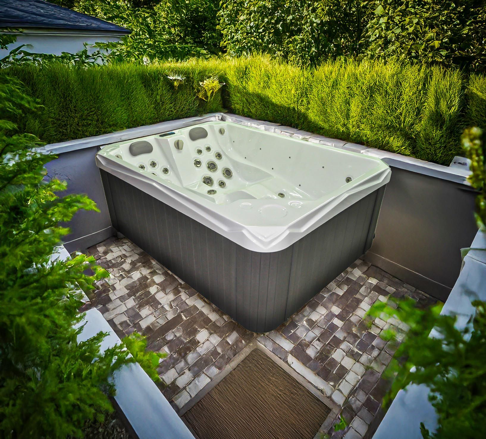 Un jacuzzi est installé dans un espace carrelé entouré de verdure. Son extérieur est gris et son intérieur est blanc.