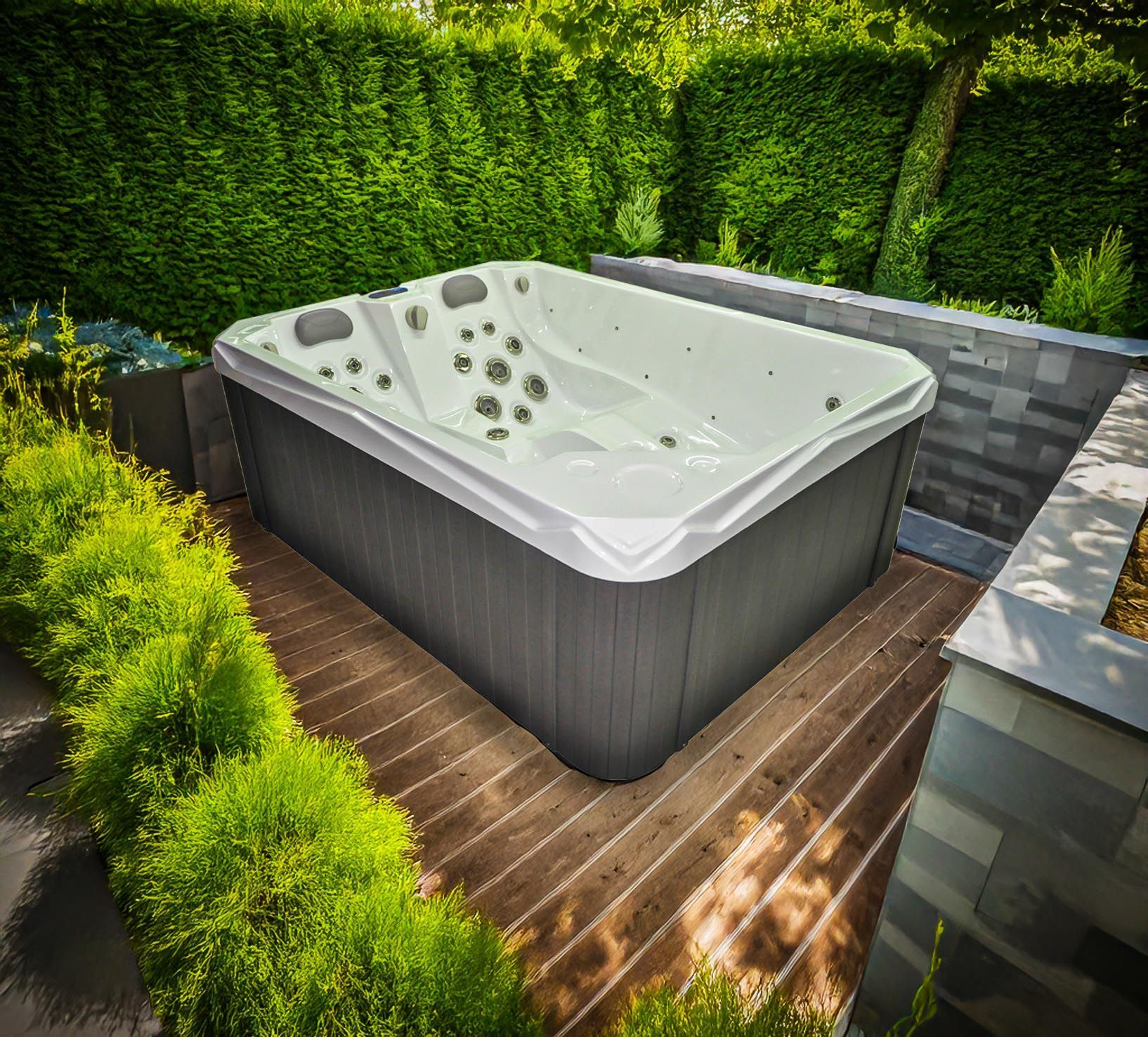 Un spa de nage rectangulaire avec des garnitures blanches et un revêtement gris se trouve sur une terrasse en bois entourée de verdure.