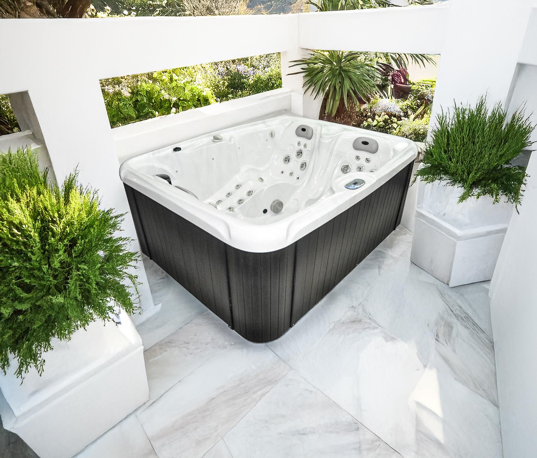 Un spa est installé sur une terrasse carrelée blanche, entourée de jardinières blanches avec des plantes vertes, sur fond de verdure.