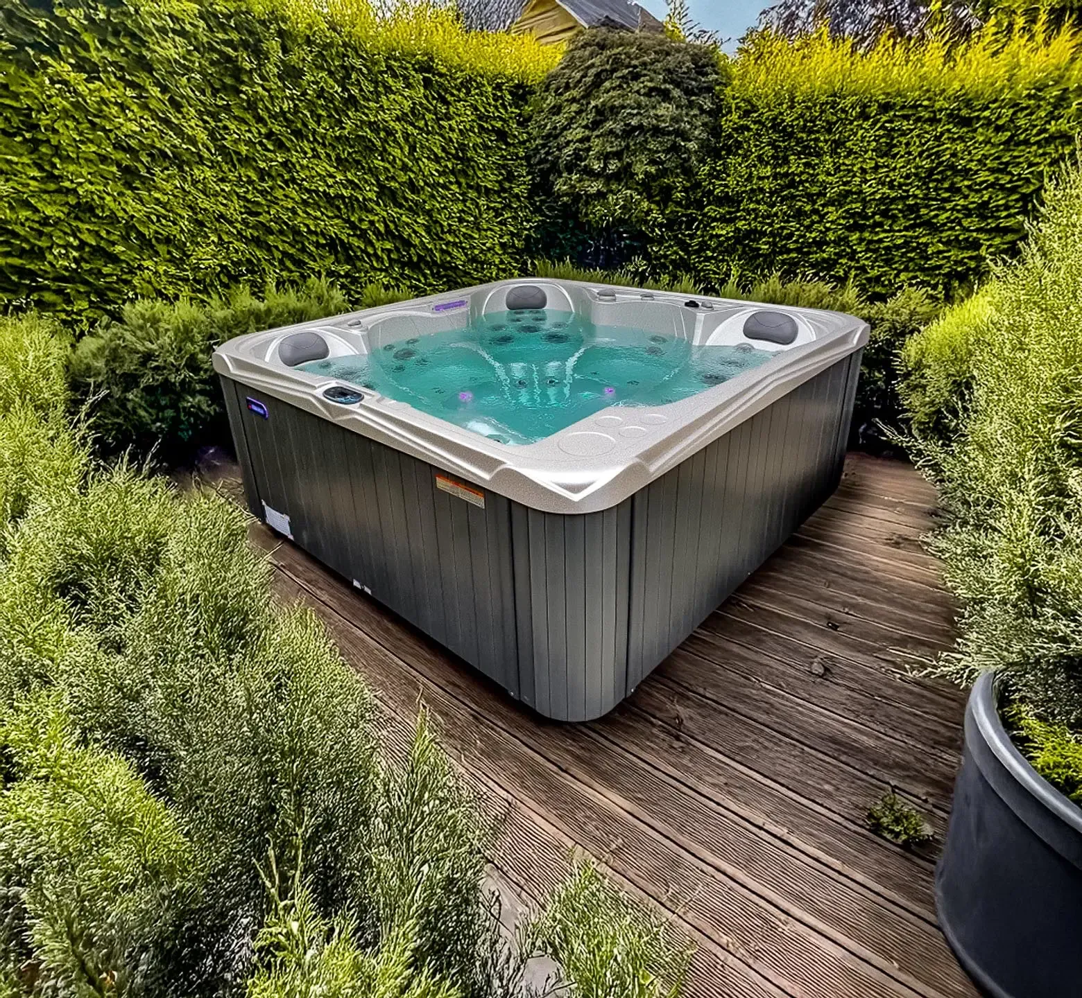 Jacuzzi entouré de verdure, installé sur une terrasse en bois. L'eau turquoise bouillonne à l'intérieur, encadrée par un extérieur gris foncé.