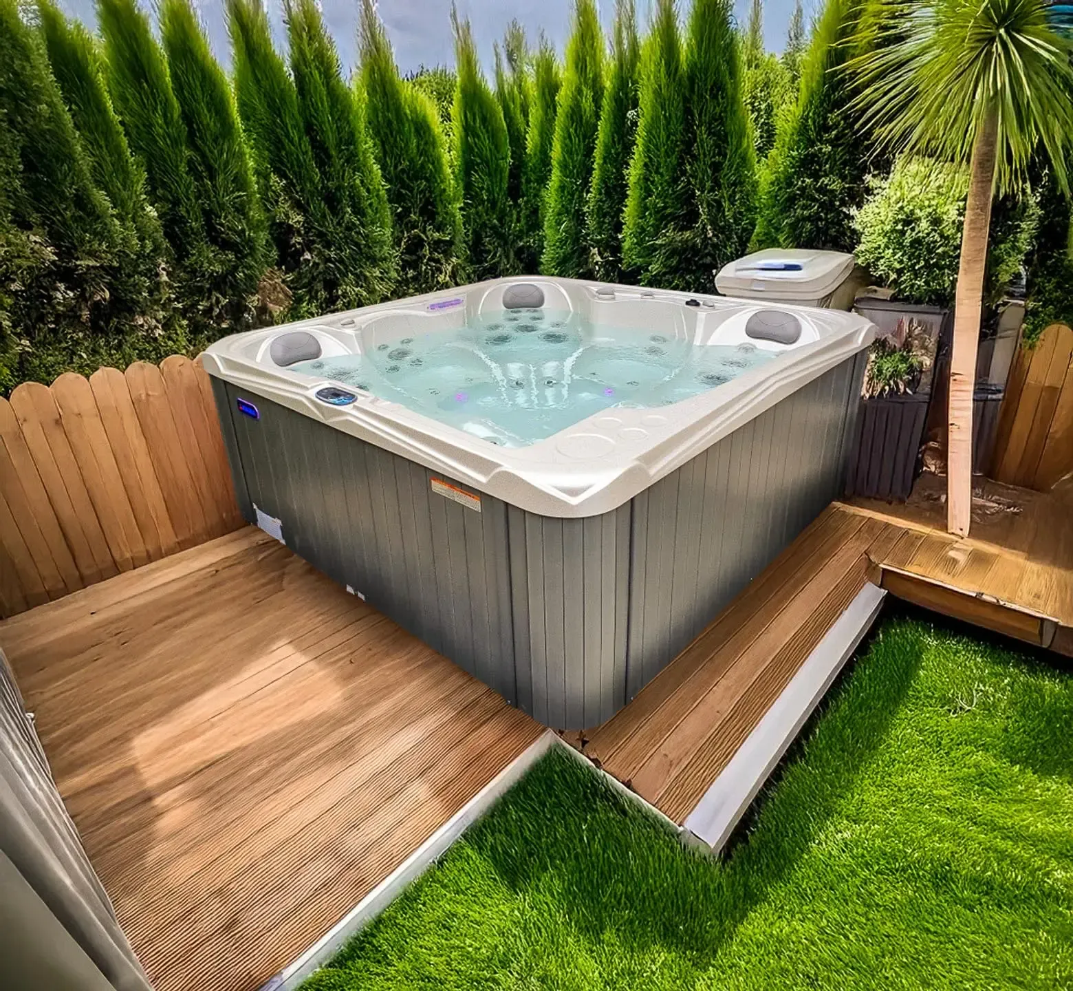 Un jacuzzi à l'eau bouillonnante est installé sur une terrasse en bois entourée de verdure. Un petit meuble blanc pour les équipements se trouve à proximité.