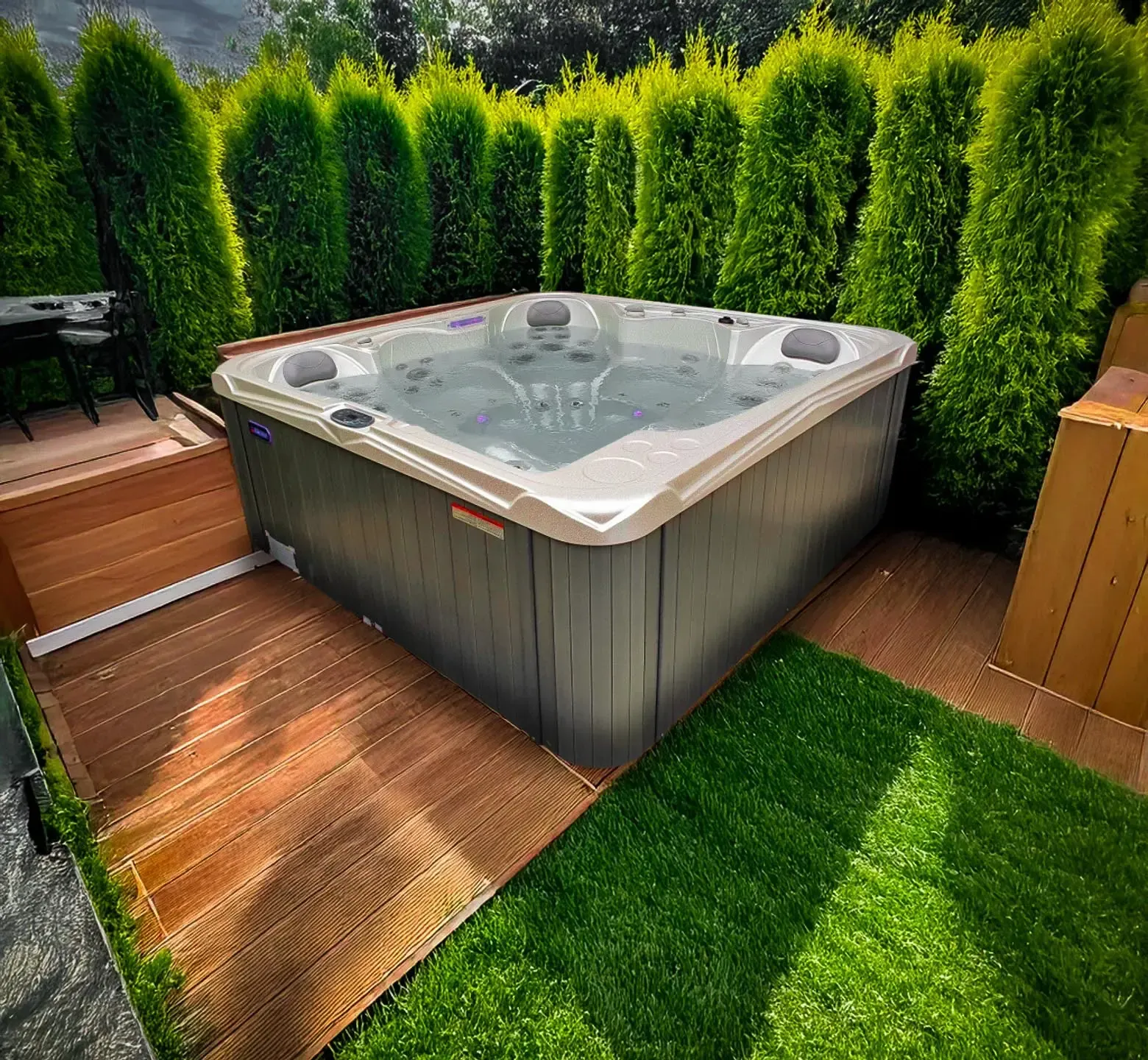 Jacuzzi entouré d'arbustes verts et d'une terrasse en bois. Le revêtement du jacuzzi est gris et l'eau est claire.