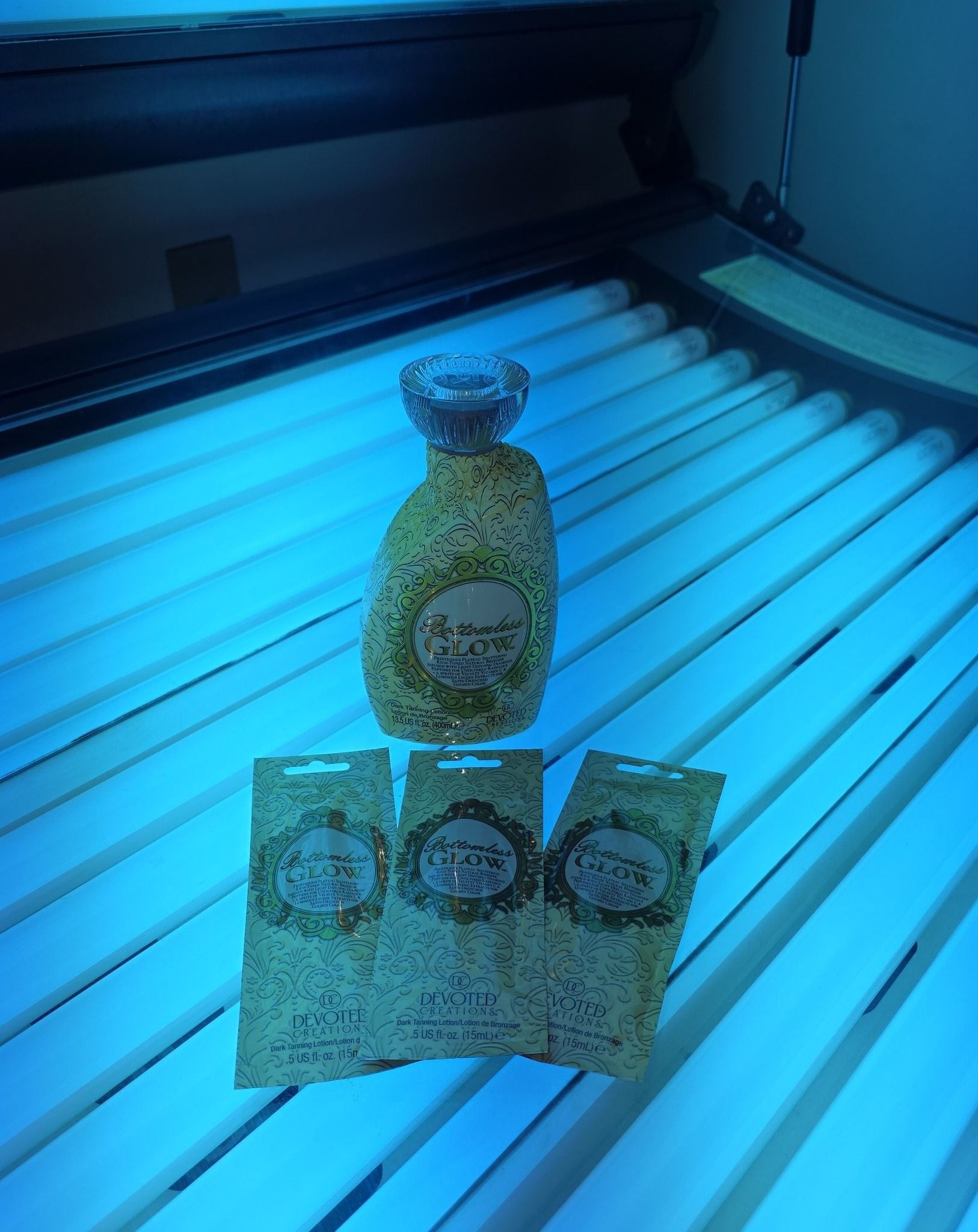 Tanning bed with a bottle and three packets of tanning lotion, all illuminated by blue UV light.
