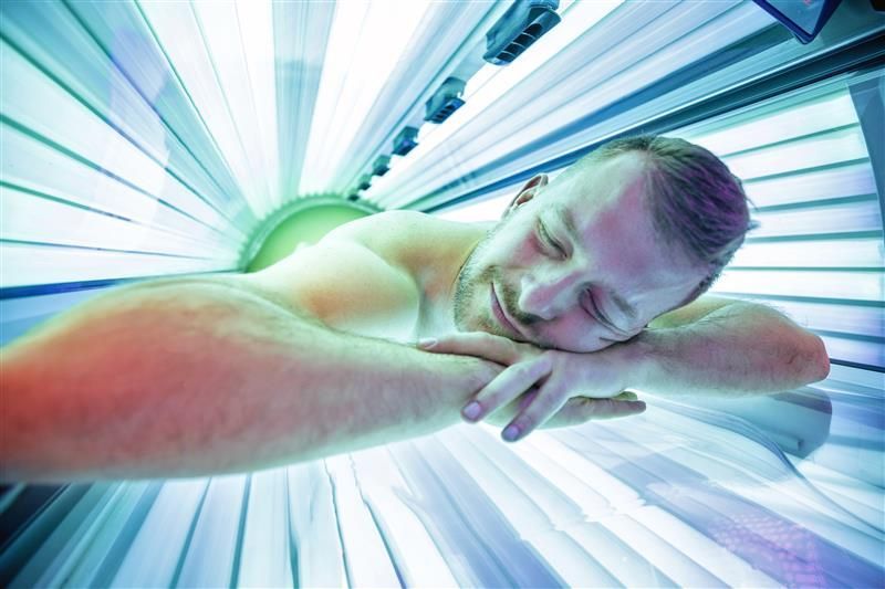 Man lying in tanning bed, eyes closed, under blue and red lights.