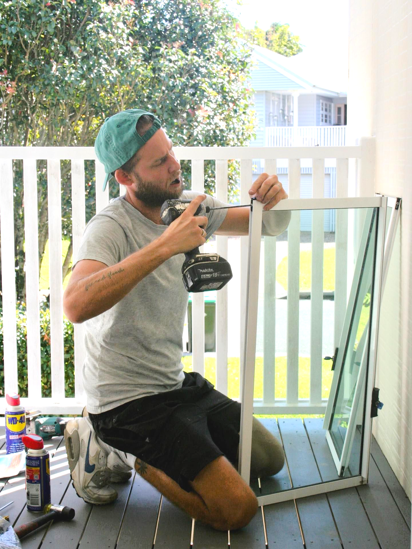 Man kneeling, using a drill on a white framed window. He wears a cap and is on a porch — Working Windows in Byron Bay, NSW