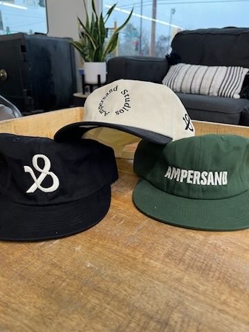 Three baseball caps on a wooden surface: black with white logo, cream with black logo, and green with white text.