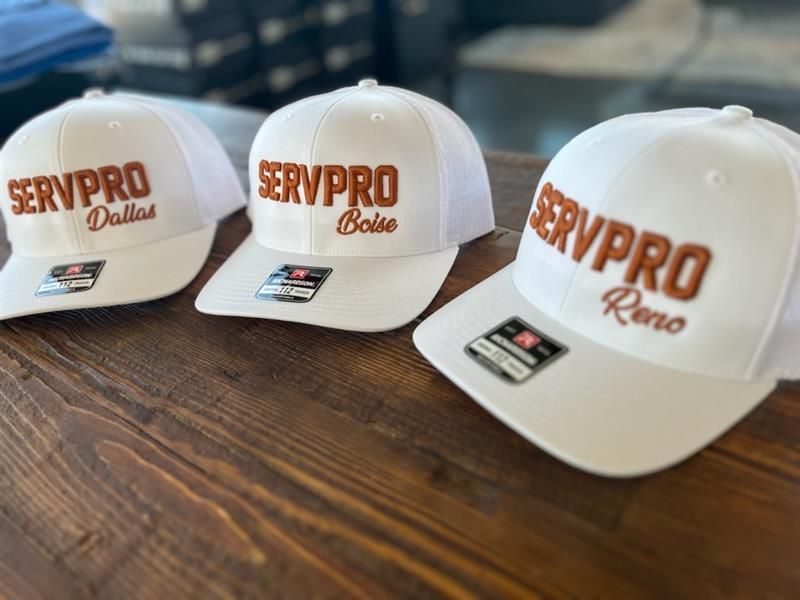 Three white SERVPPRO hats with brown lettering, each with a different city name.