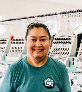 Woman smiling in a teal shirt with logo, standing in front of embroidery machines.