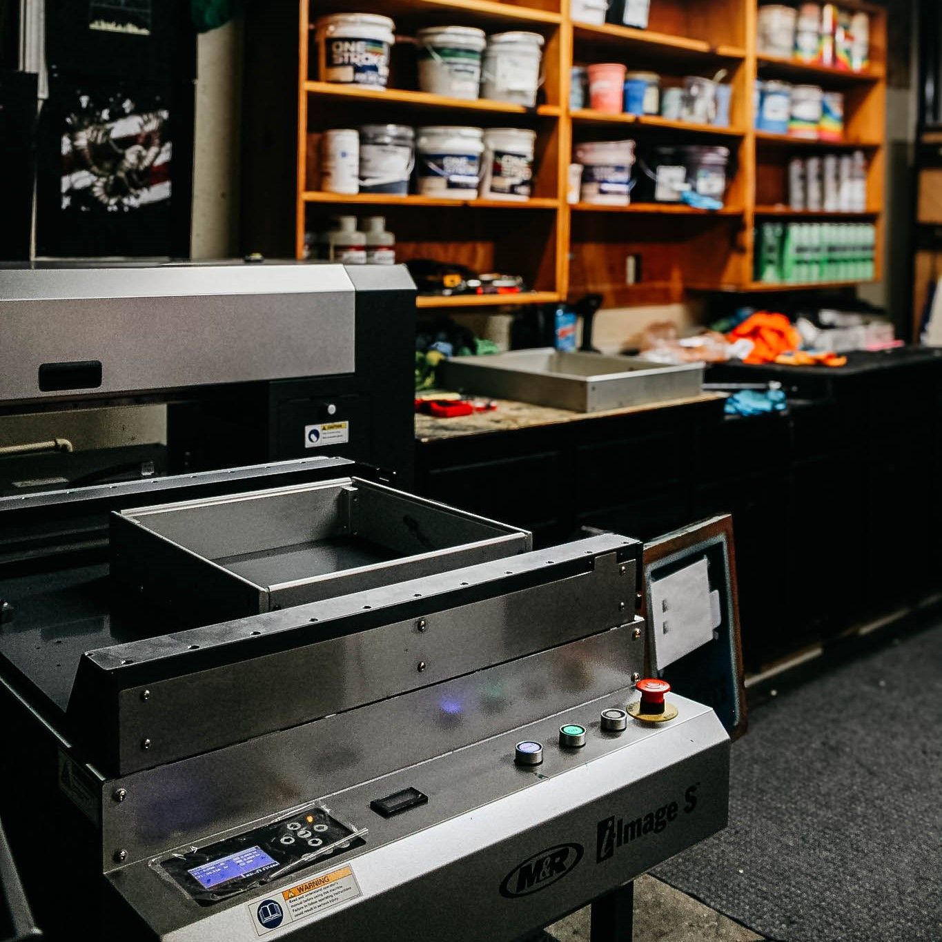 A flatbed printer in a workshop, with shelves of supplies in the background.