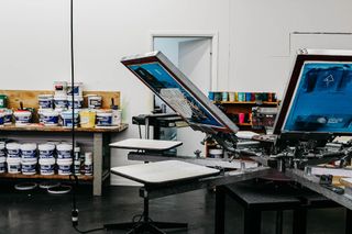 Screen printing equipment in a workshop with paint cans on a shelf in the background.