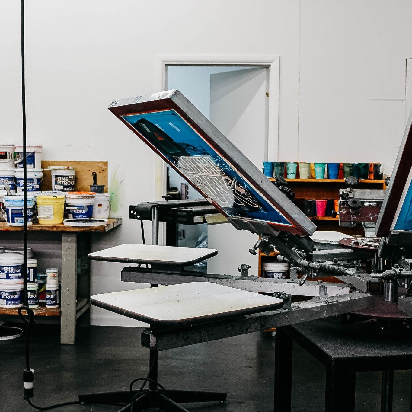 Screen printing machine in a workshop, multiple colors of ink, and a door in the background.