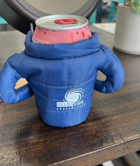 Blue puffer jacket can cooler with a pink can on a wooden table.
