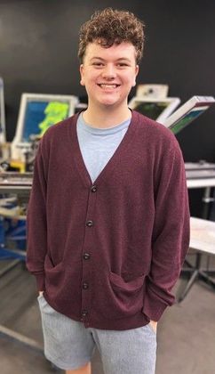 A young man with curly hair, smiling, wearing a burgundy cardigan and gray shorts, standing in a workshop.