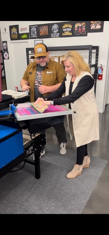 A man and woman work together at a printing station. She pulls a printed design toward her.