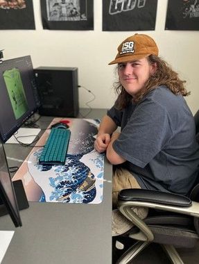 A young man with long hair and a cap sits at a desk with a computer. He smiles.