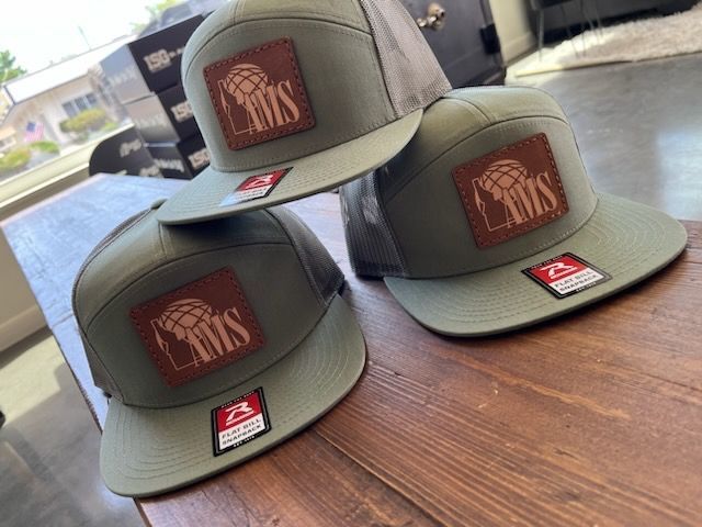 Three olive green baseball caps with brown patches and logos stacked on a wooden surface.