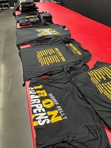 Black t-shirts with gold and white text are laid out on a red table, presumably for distribution.