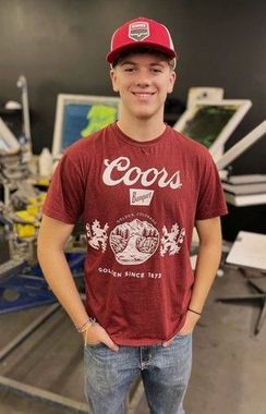 Young man in burgundy Coors t-shirt and red cap smiles, hands in pockets. Standing indoors with printing equipment.