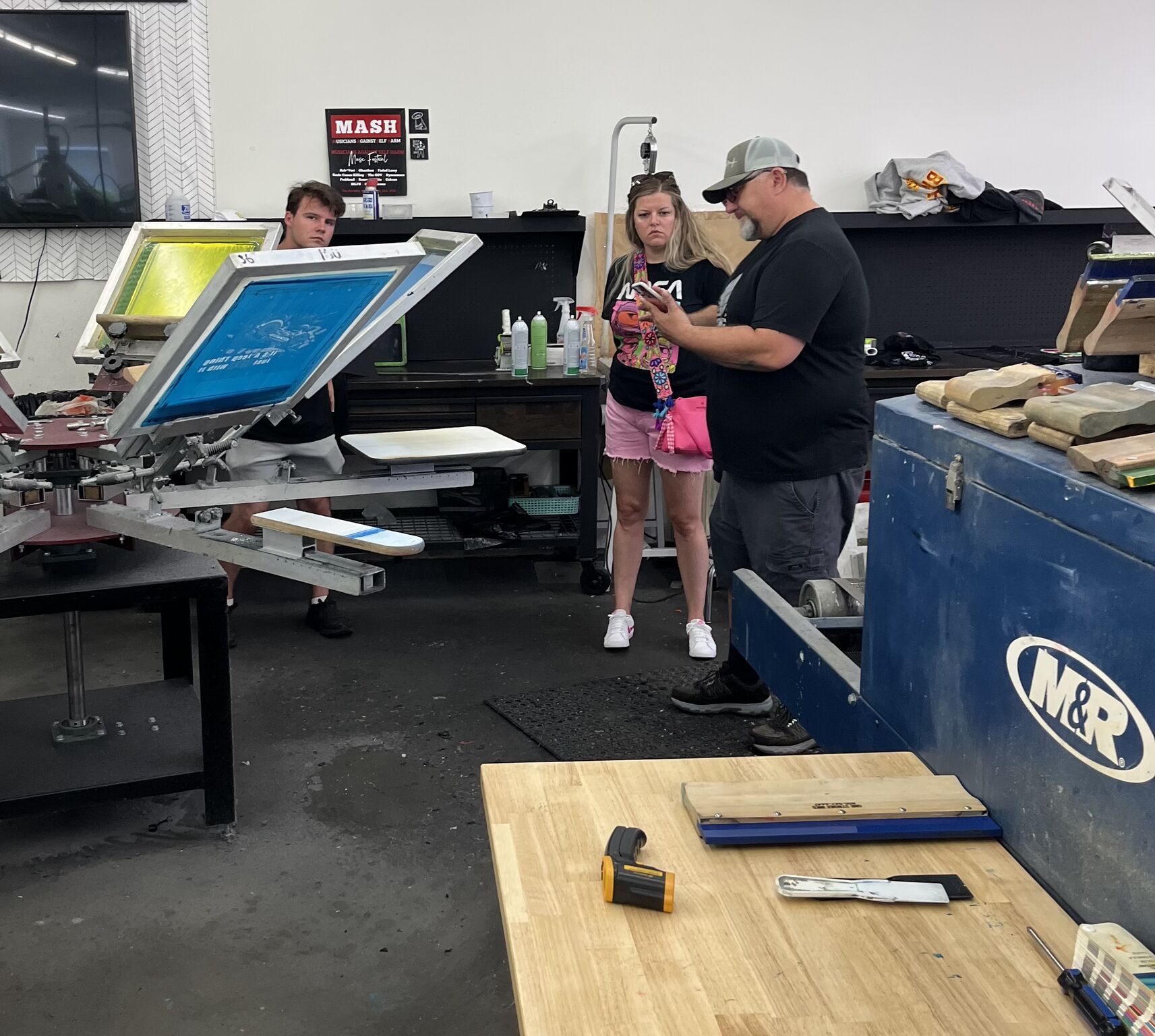 People in a screen printing shop; a man shows a phone to a woman, another watches; a printing machine in use.