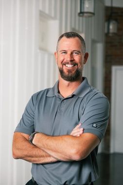 Man with arms crossed, smiling in gray polo shirt, standing in an office setting.