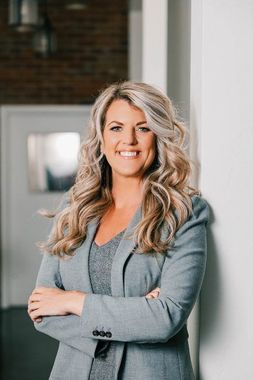 Woman with blonde wavy hair, smiling, arms crossed, wearing a gray blazer in an office setting.